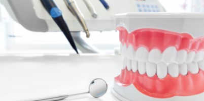 Imagem de um modelo dentário em plástico, representando uma boca com dentes, colocado sobre uma superfície branca. Ao lado do modelo, há um espelho odontológico. Ao fundo, podem ser vistos instrumentos dentários e parte de um equipamento odontológico, desfocados. A imagem evoca um ambiente de consultório odontológico, destacando a prática de cuidados dentários.