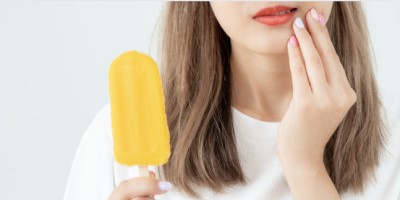 Imagem de uma mulher segurando um picolé amarelo com a mão esquerda, enquanto a mão direita toca levemente sua bochecha. A expressão facial sugere que ela está sentindo desconforto nos dentes devido ao frio do picolé. A imagem destaca o tema da sensibilidade dental.
