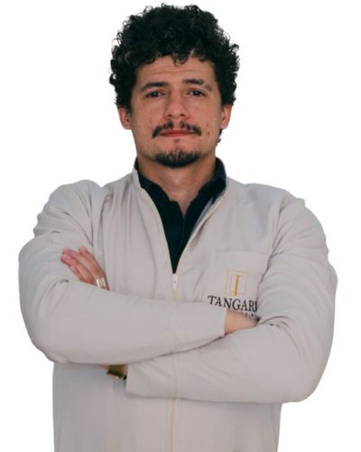 Imagem da Doutor Syrlan, ele tem cabelos pretos encaracolados e usa cavanhaque, vestindo um jaleco branco com o logotipo da Tangari Odontologia no lado esquerdo. Ele está com os braços cruzados e olha diretamente para a câmera, transmitindo uma expressão de profissionalismo.
