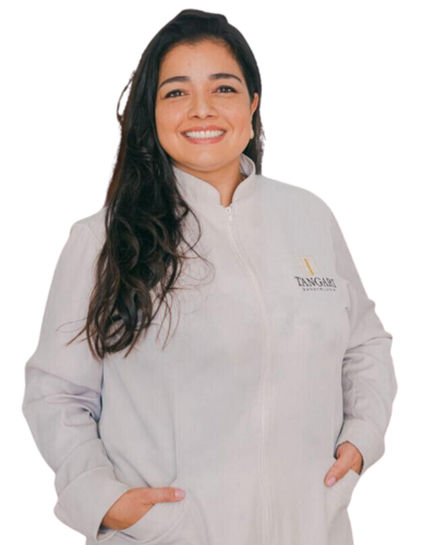 Imagem da Doutora Agnes, de cabelos longos e escuros, vestindo um jaleco branco com o logotipo da Tangari Odontologia no lado esquerdo. Ela está com as mãos nos bolsos do jaleco e olha diretamente para a câmera, transmitindo uma expressão de simpatia e profissionalismo.