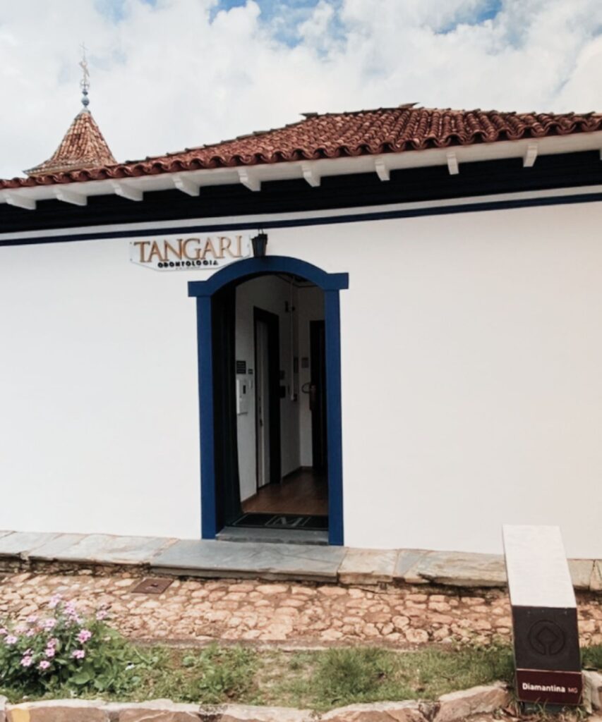A imagem mostra a fachada da clínica Tangari Odontologia. A entrada é marcada por uma porta com moldura azul, e o let TANGARI Odontologia. O edifício tem telhas de cerâmica e um estilo colonial, com paredes brancas e um adorável jardim em frente. Uma placa em madeira com o nome do consultório é visível à direita da entrada. O consultório parece estar localizado em um ambiente histórico, refletido pela arquitetura da edificação.