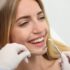 Uma mulher sorridente está em uma consulta odontológica. Ela está sentada na cadeira do dentista, enquanto um profissional de saúde, usando luvas, segura instrumentos odontológicos próximos à sua boca. Ela parece estar confortável e feliz com o atendimento.