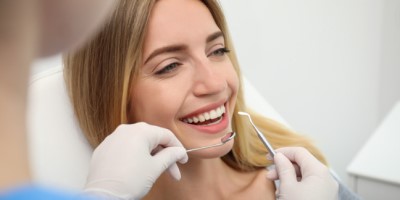 Uma mulher sorridente está em uma consulta odontológica. Ela está sentada na cadeira do dentista, enquanto um profissional de saúde, usando luvas, segura instrumentos odontológicos próximos à sua boca. Ela parece estar confortável e feliz com o atendimento.