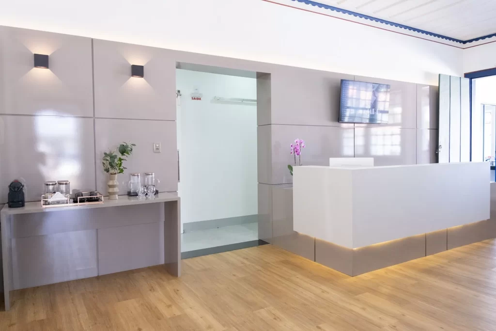 A imagem mostra a recepção da clínica Tangari odontologia. O ambiente é bem iluminado, com paredes e móveis em tons claros. À esquerda, há uma mesa com uma máquina de café, copos, recipientes decorativos e uma planta. No centro, há uma porta aberta que leva a outra sala, com uma placa indicando "SAÍDA". À direita, um balcão de recepção branco com iluminação indireta na base. Sobre o balcão, há um vaso com uma orquídea rosa. Na parede ao lado do balcão, uma televisão está montada. O chão é de madeira clara, complementando o ambiente acolhedor e sofisticado.